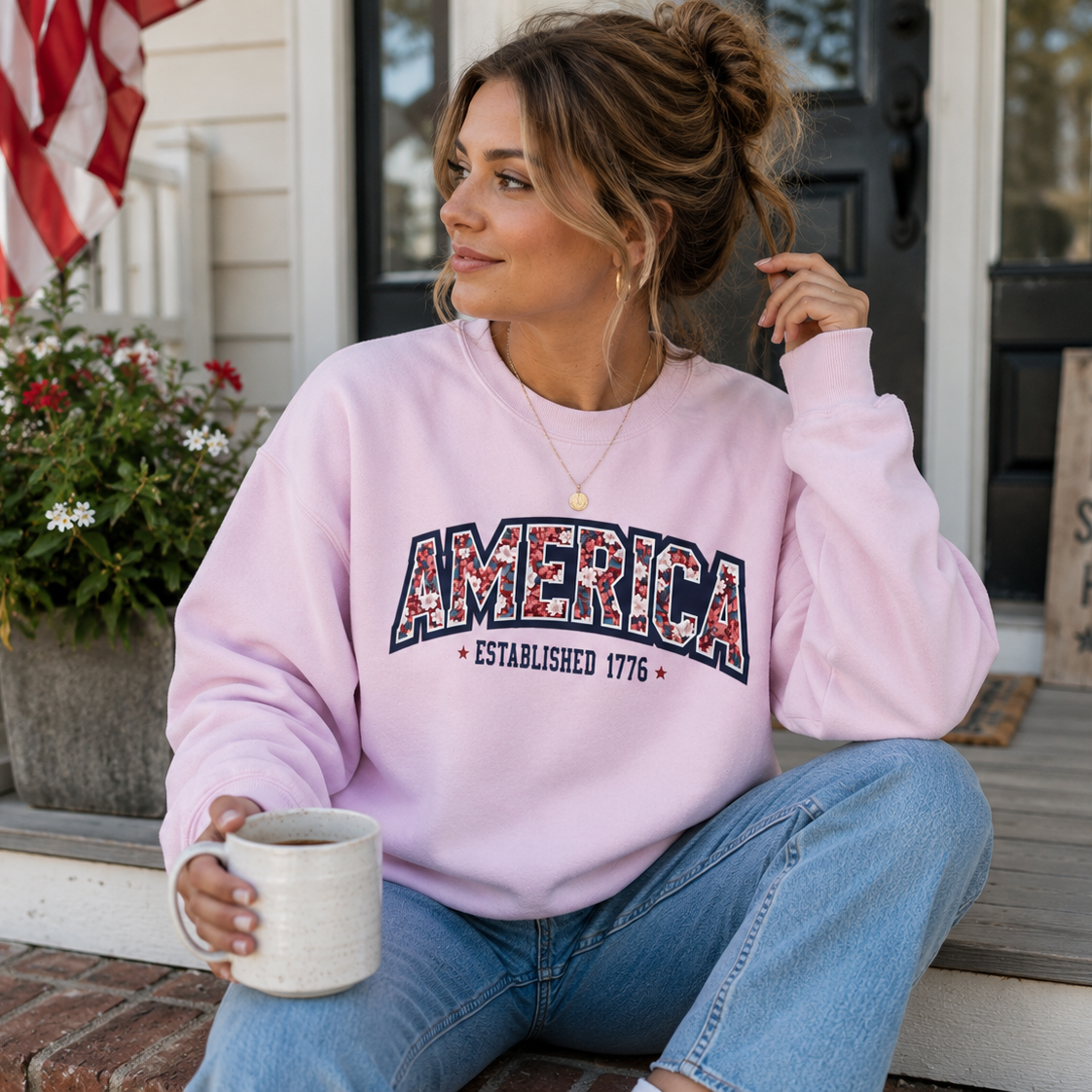 Woman wearing a pink sweatshirt with 'AMERICA' text, sitting on a porch holding a mug.