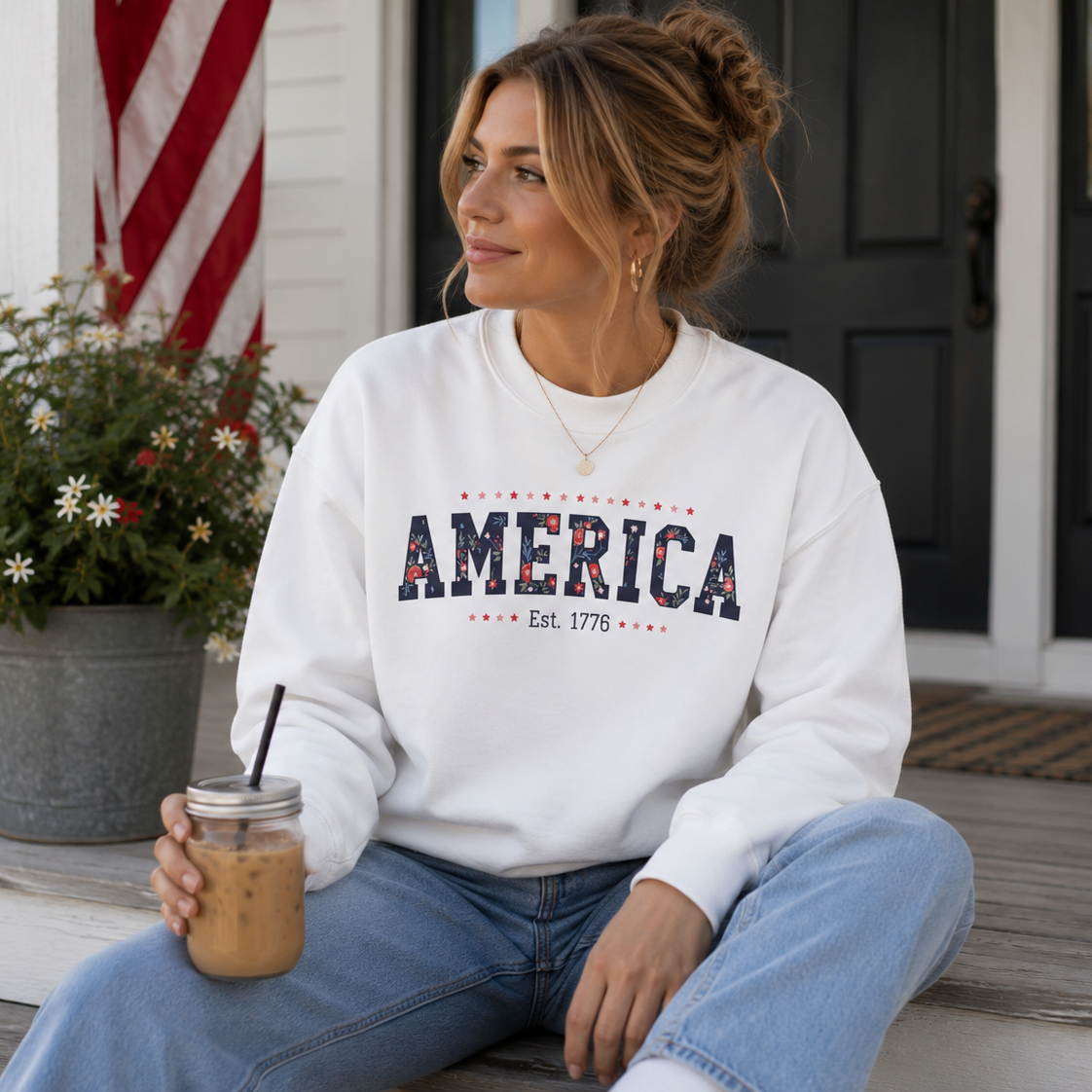 Woman sitting on a porch holding a drink, wearing a white sweatshirt with 'AMERICA' text.