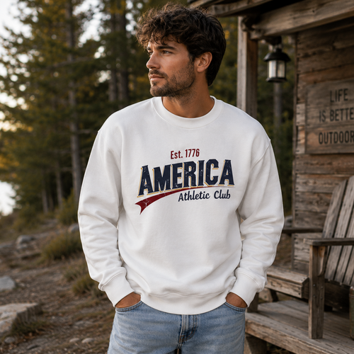 Man wearing a 'America Athletic Club' sweatshirt by a wooden cabin with a lake in the background.